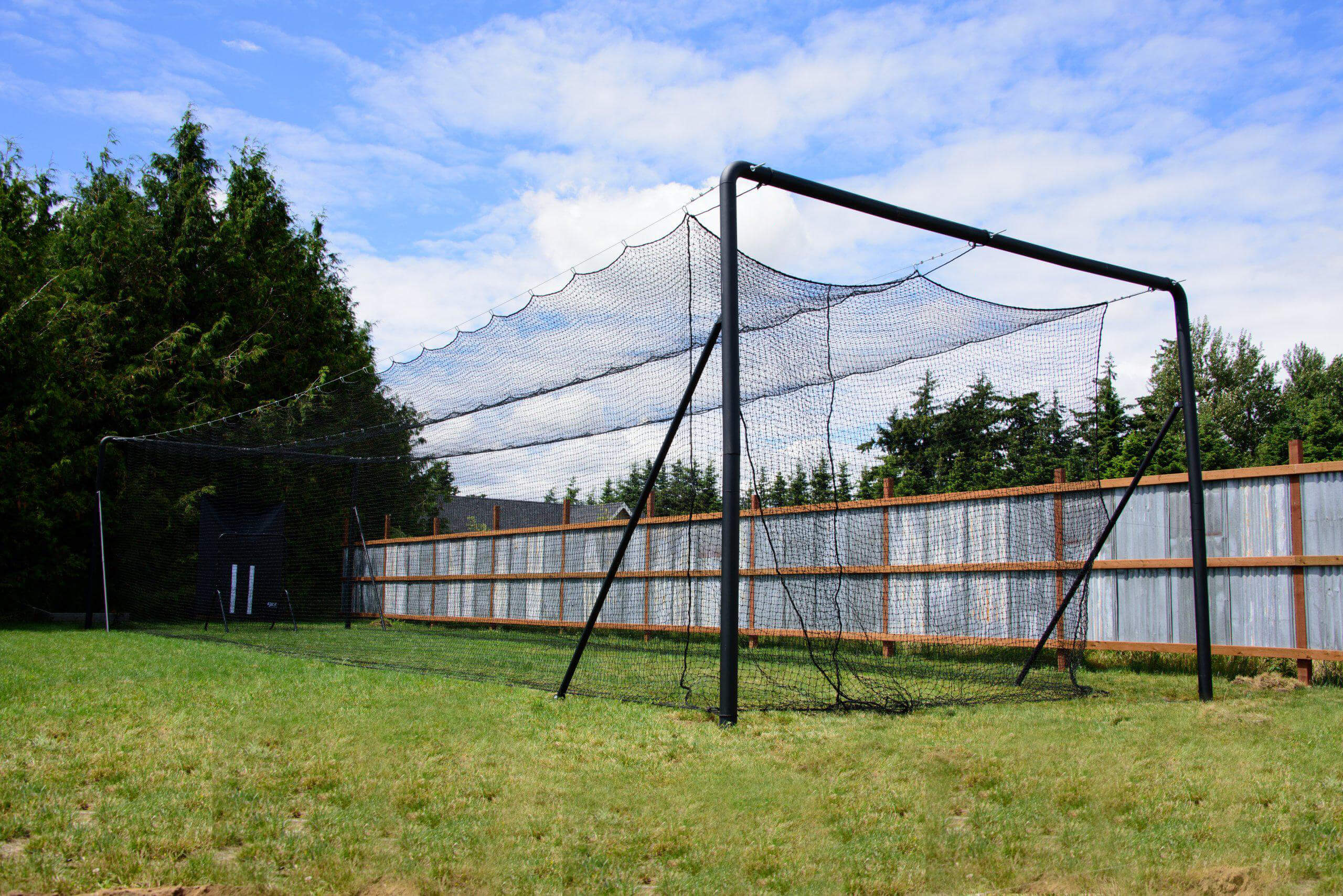 Iron Horse batting cage in backyard with fence, grass, trees, and blue skies