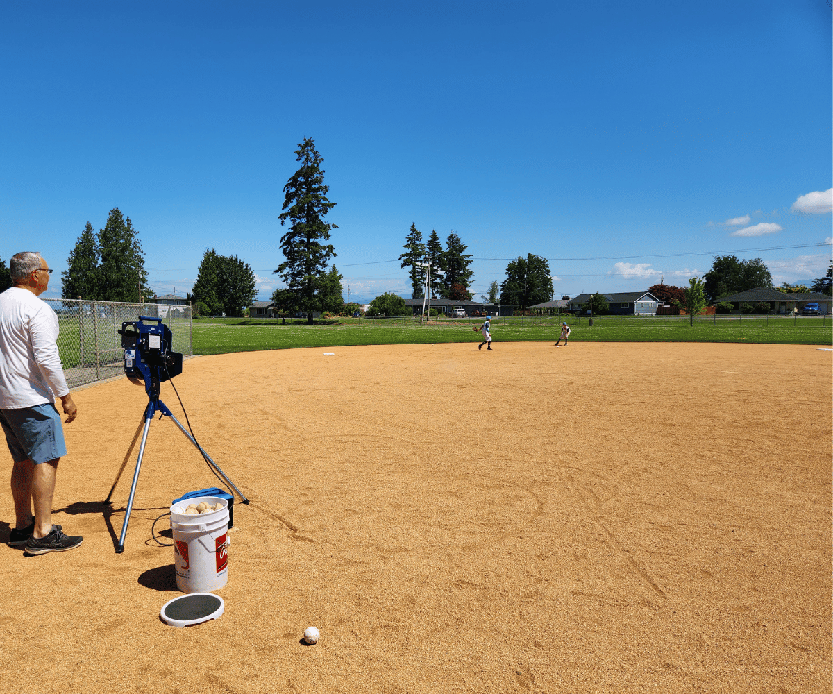 Man using a Bata 1 pitching machine to throw grounders to youth players practicing fielding