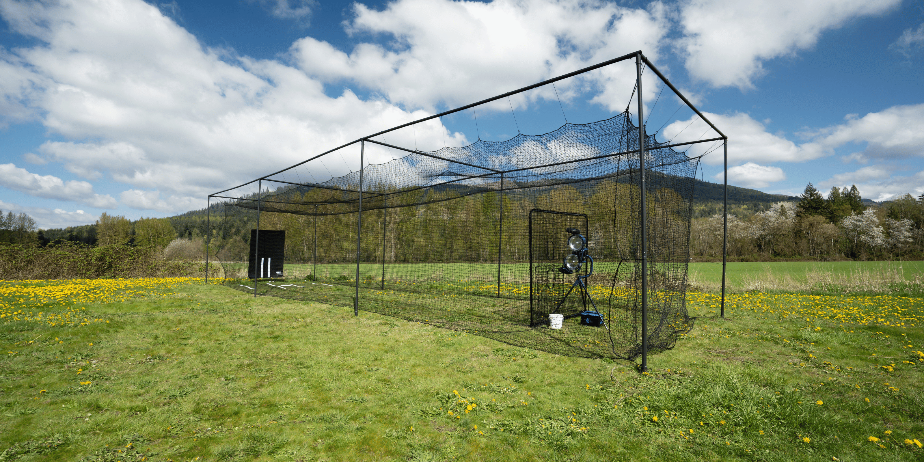 Thumper batting cage with vinyl backdrop, pitching machine, screen, and mat on top of green grass with blue skies