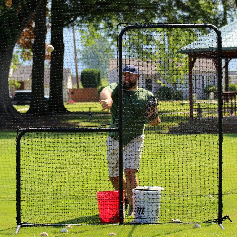 Man using the Standard Baseball Protective L-Screen while pitching with green grass behind and trees